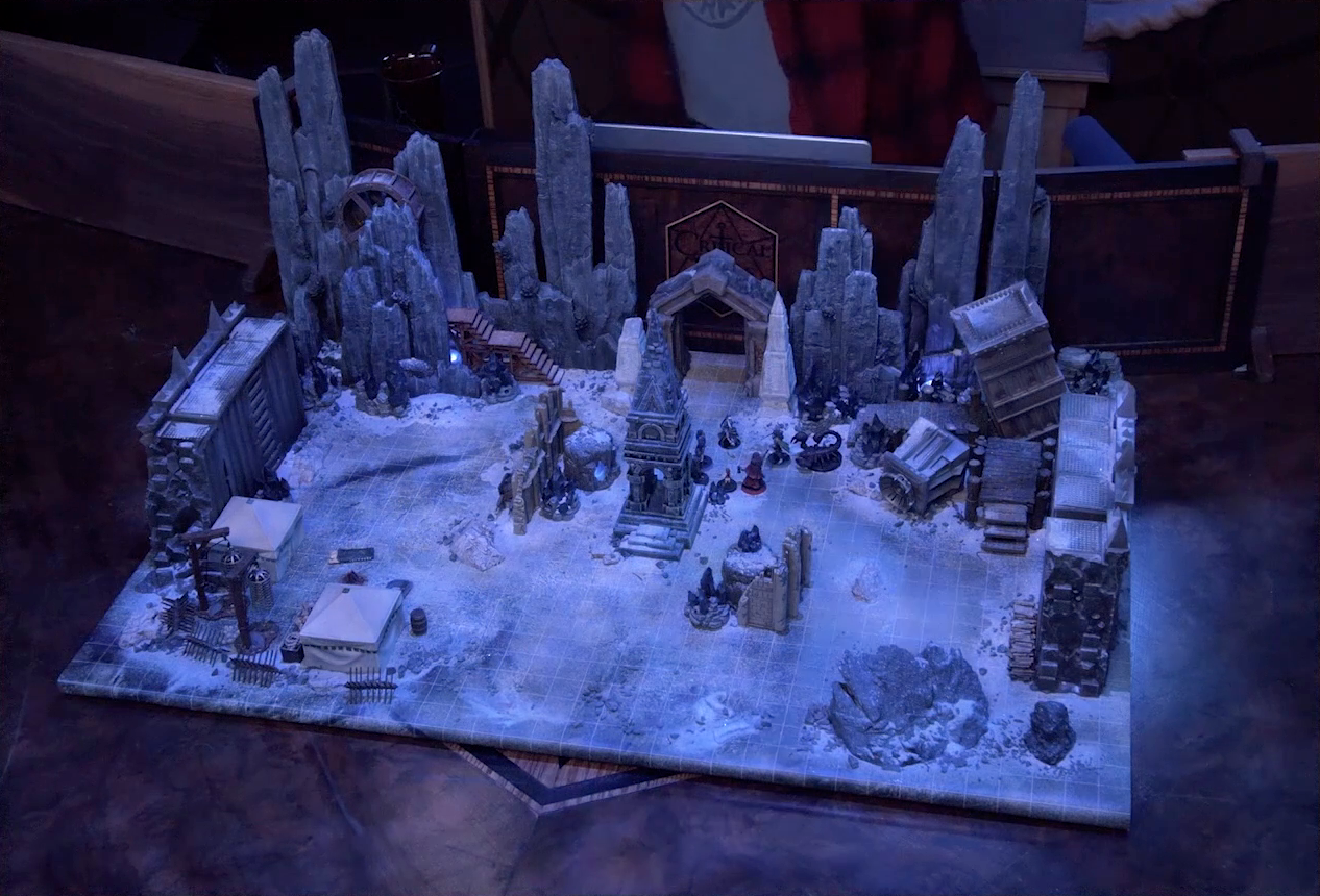 A battlemap of stony desert ruins under the cover of night. A short tower stands in the middle of the battlemap among fallen walls and collapsed towers. By the tower stand Julien, Thaisha, Occtis, Vaelus, Gaya Seremai, and Dr. Talter, as well as a giant scorpion behind Julien. A camp with two square tents, palisades, and hanging iron cages is off to the side. A gate is at the rear of the battlemap, with tall jutting rocks on either side. Some wooden stairs lead up these rocks towards a large wooden wheel. Brennan is in the background behind the DM screen. He wears a New York Public Library T-shirt under a flannel button-up.