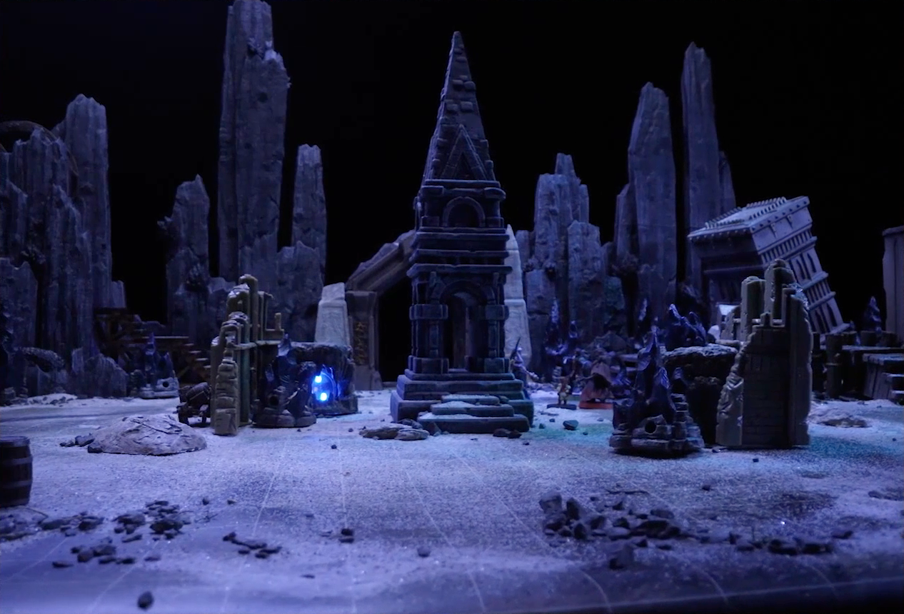 A battlemap of stony desert ruins under the cover of night. A short tower stands in the middle of the battlemap among fallen walls and collapsed towers. Vaelus, Dr. Talter, Thaisha, and a giant scorpion stand off to the side. On the other side of the tower is a blue glowing crystal in a stone enclosure. A gate is at the rear of the battlemap, with tall jutting rocks on either side. Some wooden stairs lead up these rocks towards a large wooden wheel.