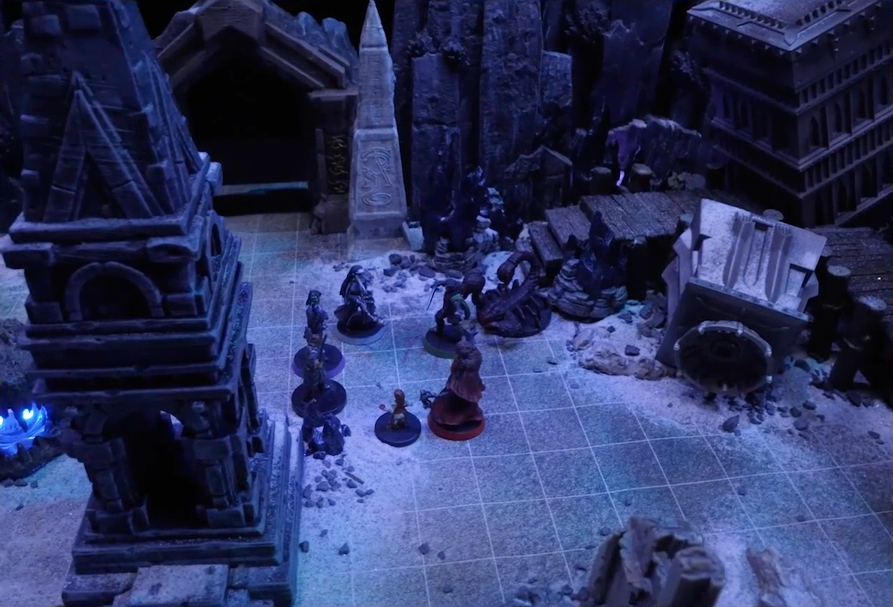 A battlemap of stony desert ruins under the cover of night focused on Occtis, Gaya Seremai, Dr. Talter, Thaisha, Julien, and Vaelus standing in a circle, with a giant scorpion directly behind Julien. On one side of the party is a short tower and a blue glowing crystal in a stone enclosure. On the other side is a collapsed ornate tower with a raised wooden boardwalk at the base of it. A gate is at the rear of the battlemap, with tall jutting rocks on either side. A gray obelisk stands to the right of the gate, a large glyph carved near its base.