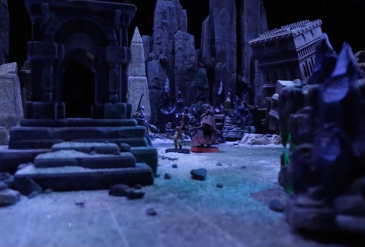 A battlemap of stony desert ruins under the cover of night focused on Occtis, Gaya Seremai, Dr. Talter, Thaisha, Julien, and Vaelus standing in a circle, with a giant scorpion directly behind Julien. On one side of the party is a short tower. On the other side is a collapsed ornate tower with a raised wooden boardwalk at the base of it. In the foreground is a green glowing crystal in a stone enclosure. A gate is at the rear of the battlemap, with tall jutting rocks on either side. A gray obelisk stands to the right of the gate, a large glyph carved near its base.