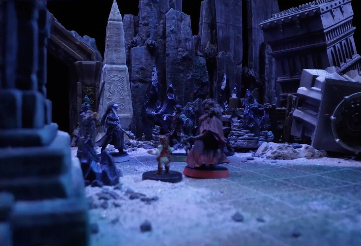 A battlemap of stony desert ruins under the cover of night focused on Occtis, Gaya Seremai, Dr. Talter, Thaisha, Julien, and Vaelus standing in a circle, with a giant scorpion directly behind Julien. On one side of the party is a short tower. On the other side is a collapsed ornate tower with a raised wooden boardwalk at the base of it. A gate is at the rear of the battlemap, with tall jutting rocks on either side. A gray obelisk stands to the right of the gate, a large glyph carved near its base.