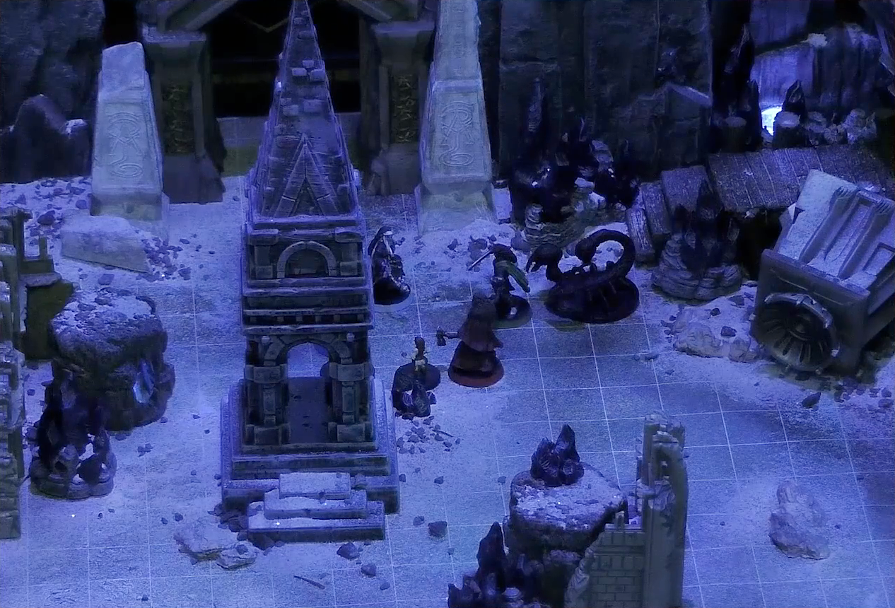 A battlemap of stony desert ruins under the cover of night focused on Occtis, Dr. Talter, Thaisha, Julien, and Vaelus standing in a circle, with a giant scorpion directly behind Julien. On one side of the party is a short tower and a blue glowing crystal in a stone enclosure. On the other side is a collapsed ornate tower with a raised wooden boardwalk at the base of it. A gate is at the rear of the battlemap, with tall jutting rocks on either side. Two gray obelisks stand to the sides of the gate, large glyphs carved near their bases.