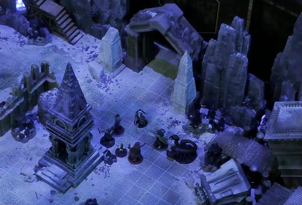 A battlemap of stony desert ruins under the cover of night focused on Occtis, Gaya Seremai, Dr. Talter, Thaisha, Julien, and Vaelus standing in a circle, with a giant scorpion directly behind Julien. On one side of the party is a short tower and a blue glowing crystal in a stone enclosure. On the other side is a collapsed ornate tower with a raised wooden boardwalk at the base of it. A gate is at the rear of the battlemap, with tall jutting rocks on either side. Two gray obelisks stand to the sides of the gate, large glyphs carved near their bases.