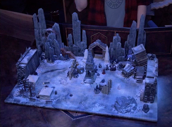 A battlemap of stony desert ruins under the cover of night. A short tower stands in the middle of the battlemap among fallen walls and collapsed towers. By the tower stand Julien, Thaisha, Occtis, Vaelus, Gaya Seremai, and Dr. Talter, as well as a giant scorpion behind Julien. Dr. Talter is much further away on the other side of the tower. A camp with two square tents, palisades, and hanging iron cages is off to the side. A gate is at the rear of the battlemap, with tall jutting rocks on either side. Some wooden stairs lead up these rocks towards a large wooden wheel.