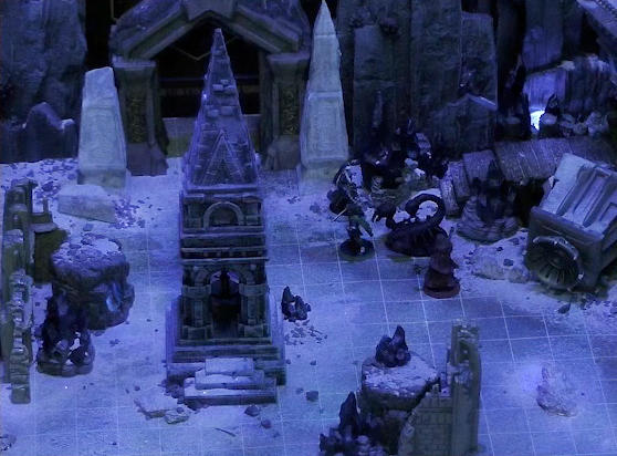 A battlemap of stony desert ruins under the cover of night focused on Gaya Seremai, Thaisha, Julien, and Vaelus facing off against a giant scorpion. On one side of the party is a short tower and a blue glowing crystal in a stone enclosure. Dr. Talter is in the tower. On the other side is a collapsed ornate tower with a raised wooden boardwalk at the base of it. A gate is at the rear of the battlemap, with tall jutting rocks on either side. Two gray obelisks stand to the sides of the gate, large glyphs carved near their bases.