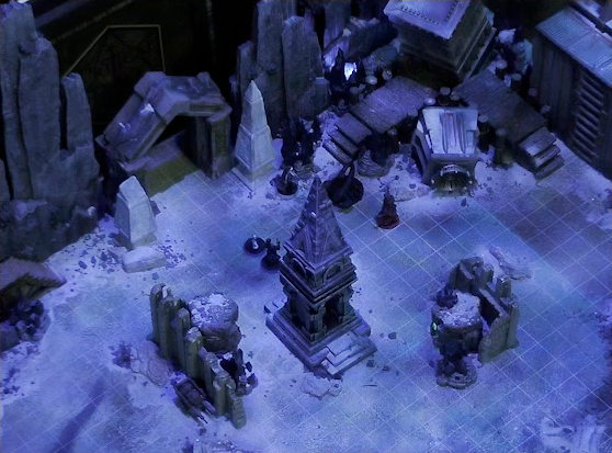 A battlemap of stony desert ruins under the cover of night focused on Gaya Seremai, Thaisha, Julien, and Vaelus facing off against a giant scorpion. On one side of the party is a short tower, with Occtis and Pin behind it. On the other side is a collapsed ornate tower with a raised wooden boardwalk at the base of it. A gate is at the rear of the battlemap, with tall jutting rocks on either side. Two gray obelisks stand to the sides of the gate, large glyphs carved near their bases.