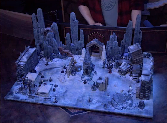 A battlemap of stony desert ruins under the cover of night. The map is filled with a large number of skeletons. A short tower stands in the middle of the battlemap among fallen walls and collapsed towers. Gaya Seremai, Thaisha, Julien, and Vaelus face off against a giant scorpion. A camp with two square tents, palisades, and hanging iron cages is off to the side. A gate is at the rear of the battlemap, with tall jutting rocks on either side. Some wooden stairs lead up these rocks towards a large wooden wheel. Brennan is in the background behind the DM screen. He wears a New York Public Library T-shirt under a flannel button-up.