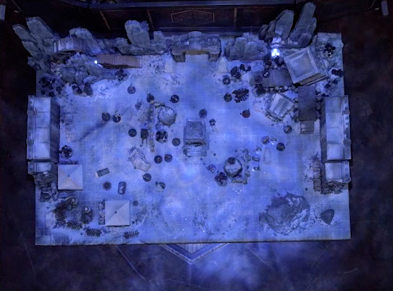 A top-down view of a battlemap of stony desert ruins under the cover of night. The map is filled with a large number of skeletons. A short tower stands in the middle of the battlemap among fallen walls and collapsed towers. Gaya Seremai, Thaisha, Julien, and Vaelus face off against a giant scorpion. A camp with two square tents, palisades, and hanging iron cages is off to the side. A gate is at the rear of the battlemap, with tall jutting rocks on either side. Some wooden stairs lead up these rocks towards a large wooden wheel.
