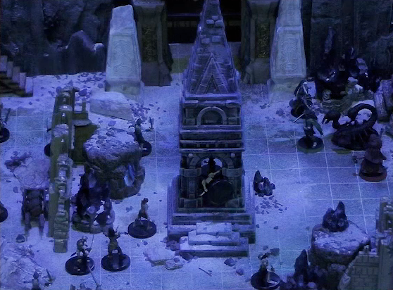 A battlemap of stony desert ruins under the cover of night. A short tower stands in the middle of the battlemap. Gaya Seremai, Thaisha, Julien, and Vaelus face off against a giant scorpion to the right of the tower. A downed skeleton is in the tower, as is Dr. Talter. Several other skeletons approach the tower. A gate is at the rear of the battlemap, with tall jutting rocks on either side. Two gray obelisks stand to the sides of the gate, large glyphs carved near their bases.