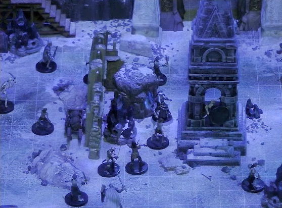 A battlemap of stony desert ruins under the cover of night focused on a short tower. Vaelus faces off against several approaching skeletons outside the tower. A downed skeleton is in the tower. In the background is visible a gate, with tall jutting rocks on either side. Two gray obelisks stand to the sides of the gate, large glyphs carved near their bases. Some wooden stairs lead up the jutting rocks to the left of the gate.
