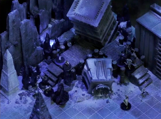 A battlemap of stony desert ruins under the cover of night focused on Pin, Dr. Talter, and Occtis facing off against a group of approaching skeletons. They stand on a raised wooden boardwalk at the base of a collapsed ornate tower. The skeletons climb short stairs at the edge of the boardwalk. A giant scorpion is also at the base of the boardwalk. Barely visible in the foreground is the tip of a stone tower, and off to the side is a gray obelisk, a large glyph carved near its base.