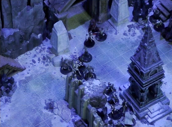 A battlemap of stony desert ruins under the cover of night focused on a short tower. Vaelus faces off against several approaching skeletons outside the tower. Thaisha and Julien face off against more skeletons near a gate at the back of the battlemap. Two gray obelisks stand to the sides of the gate, large glyphs carved near their bases.