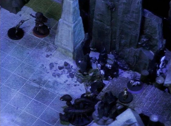 A battlemap of stony desert ruins under the cover of night focused on Pin and Dr. Talter on a raised wooden boardwalk at the base of a collapsed ornate tower. Gaya Seremai is below them near a giant scorpion, facing off against a skeleton. Thaisha faces off against another skeleton by a gate. Off to the right of the gate is a gray obelisk, a large glyph carved near its base.