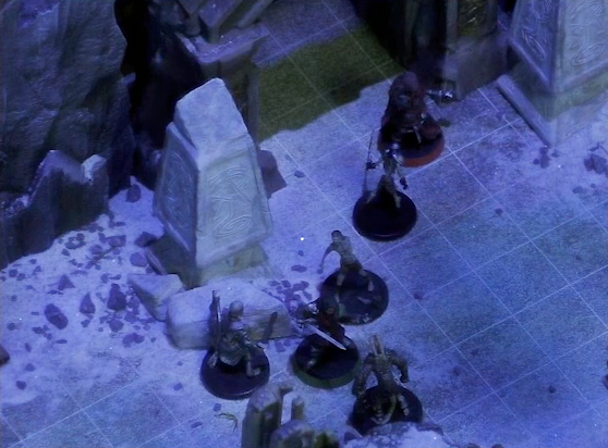 A battlemap of stony desert ruins under the cover of night focused on Thaisha and Julien facing off against four skeletons in front of a gate. Two gray obelisks stand to the sides of the gate, large glyphs carved near their bases.