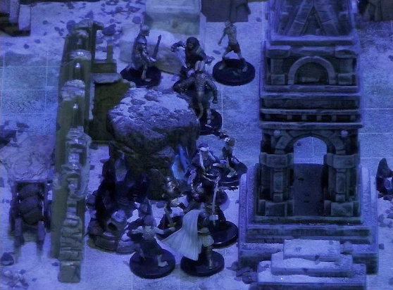 A battlemap of stony desert ruins under the cover of night focused on a short tower. Vaelus faces off against several approaching skeletons outside the tower. Thaisha and Julien face off against more skeletons near a gate at the back of the battlemap.