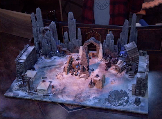 A battlemap of stony desert ruins under the cover of night. A short tower stands in the middle of the battlemap among fallen walls and collapsed towers. A soft dawn glow falls on the tower. Thaisha, Julien, Vaelus, Occtis, Gaya Seremai, and Dr. Talter face off against a number of skeletons throughout the map. A camp with two square tents, palisades, and hanging iron cages is off to the side. A gate is at the rear of the battlemap, with tall jutting rocks on either side. Some wooden stairs lead up these rocks towards a large wooden wheel. Brennan is in the background behind the DM screen. He wears a New York Public Library T-shirt under a flannel button-up.