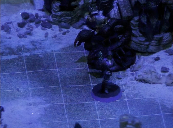 A battlemap of stony desert ruins under the cover of night focused Occtis standing in front of a giant scorpion. Behind the large black scorpion is a skeleton carrying an ax, and behind it are wooden steps leading up some rocks.