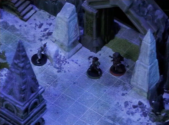 A battlemap of stony desert ruins under the cover of night focused on Thaisha and Julien facing off against a skeleton in front of a gate. Two gray obelisks stand to the sides of the gate, large glyphs carved near their bases. A short tower raises in the foreground.