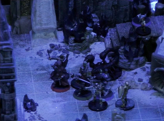 A battlemap of stony desert ruins under the cover of night focused on Thaisha, Julien, Occtis, and Gaya Seremai facing off against some skeletons and a giant scorpion. Pin and Dr. Talter stand on a raised wooden boardwalk at the base of a collapsed ornate tower. In the background is a gate, with a gray obelisk to its right side, a large glyph carved near its base.