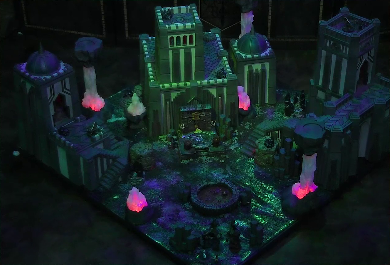 A battlemap of a walled courtyard under cover of night. Four towers stand in a row with a metal sacrificial platform bearing a pentagram sitting before a center tower. In front of the pentagram is a circular pool of dark red blood. Interspersed throughout the chamber are six red glowing crystal pillars. One ballista is on the lower platform of the left tower, and another is on top of the right tower. Gaya Seremai, Pin, Vaelus, Julien, Occtis, Dr. Talter, and Thaisha stand at the edge of the battlemap, looking towards the pool of blood.