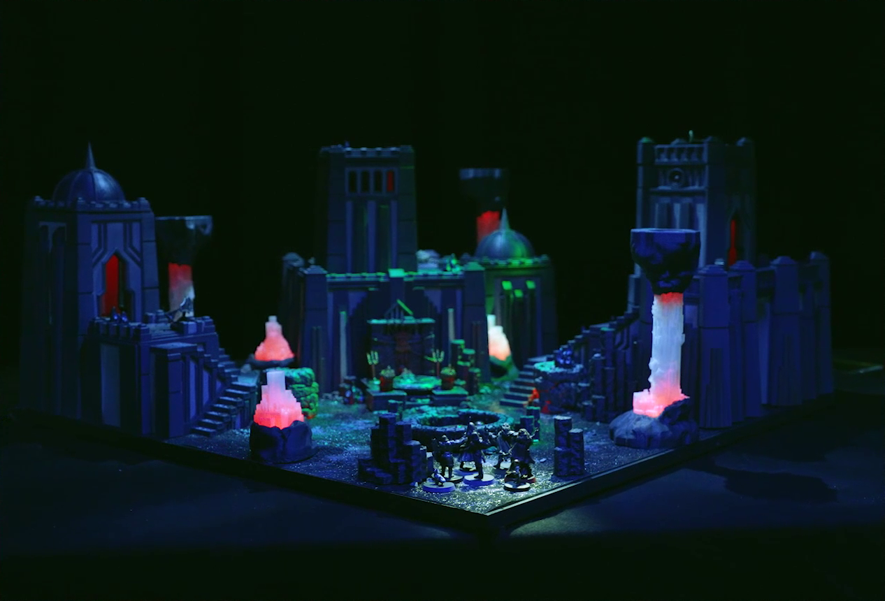 A battlemap of a walled courtyard under cover of night. Four towers stand in a row with a metal sacrificial platform bearing a pentagram sitting before a center tower. In front of the pentagram is a circular pool of dark red blood. Interspersed throughout the chamber are six red glowing crystal pillars. One ballista is on the lower platform of the left tower, and another is on top of the right tower. Gaya Seremai, Pin, Vaelus, Julien, Occtis, Dr. Talter, and Thaisha stand at the edge of the battlemap, looking towards the pool of blood.