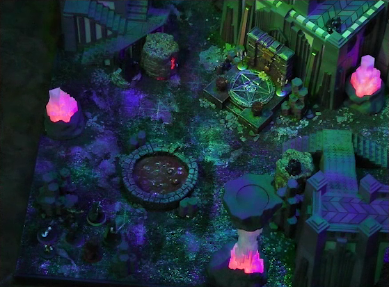 A battlemap of a walled courtyard under cover of night. Four towers stand in a row with a metal sacrificial platform bearing a pentagram sitting before a center tower. Two candelabras are at the corners of the platform. In front of the pentagram is a circular pool of dark red blood. Interspersed throughout the chamber are at least three red glowing crystal pillars. Gaya Seremai, Pin, Vaelus, Julien, Occtis, Dr. Talter, and Thaisha stand at the edge of the battlemap, looking towards the pool of blood.