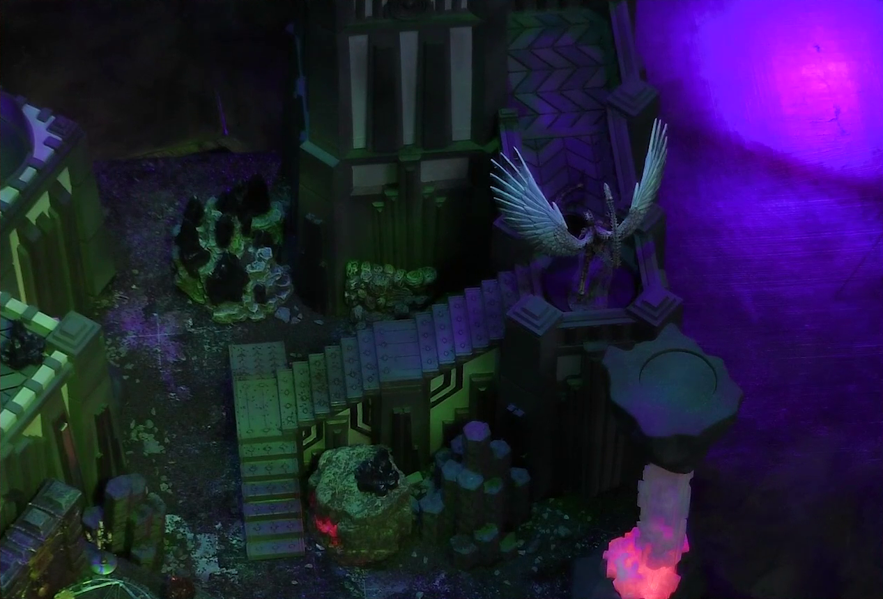 A battlemap of a walled courtyard under cover of night focused on the white-winged and scorpion-tailed Tertia Tachonis on a stone wall at the top of a long flight of steps. Her wings are outspread and her skeletal scorpion tail arches above her head. At her feet is the downed body of Gaya Seremai. A red glowing crystal pillar is at the base of the stairs. Off to the side a couple towers are visible, with a metal platform at the base of one and a candelabra at its corner.