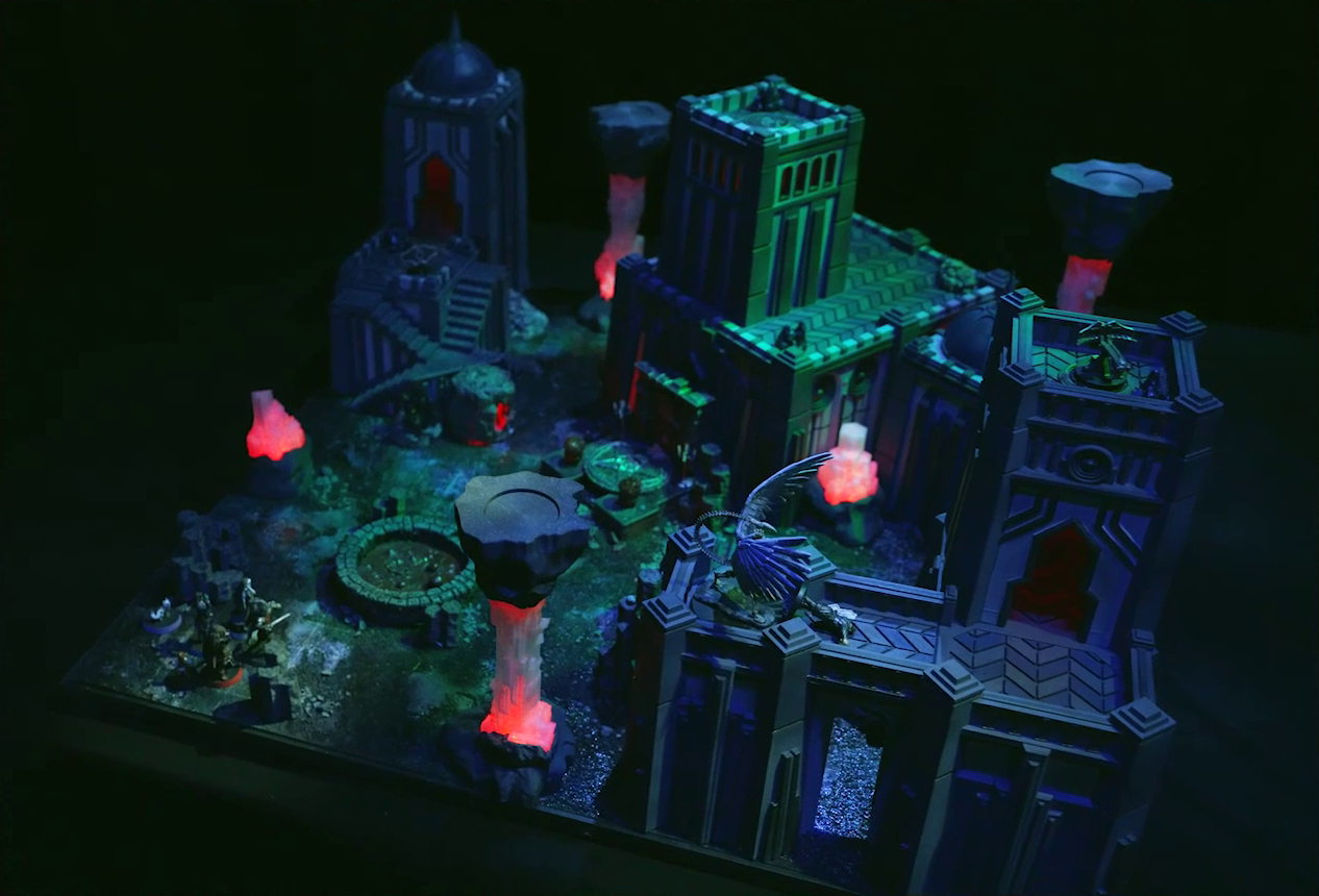 A battlemap of a walled courtyard under cover of night. Four towers stand in a row with a metal sacrificial platform bearing a pentagram sitting before a center tower. The white-winged and scorpion-tailed Tertia Tachonis is on top of a wall off the right-most tower. At her feet is the downed body of Gaya Seremai. In front of the pentagram is a circular pool of dark red blood. Interspersed throughout the chamber are six red glowing crystal pillars. One ballista is on the lower platform of the left tower, and another is on top of the right tower. Pin, Vaelus, Julien, Occtis, Dr. Talter, and Thaisha stand at the edge of the battlemap, looking towards the pool of blood.