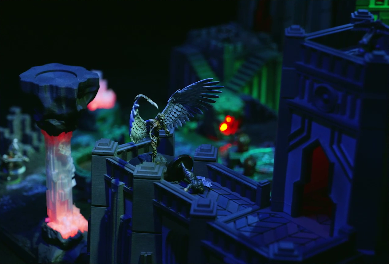 A battlemap of a walled courtyard under cover of night focused on the white-winged and scorpion-tailed Tertia Tachonis on a stone wall standing over the body of Gaya Seremai. Tertia’s body is gaunt and zombie-like, dirty yellow clothes laying loosely across her frame. She leans forward with clawed hands, her wings outstretched and her skeletal tail arched above her head. Behind Tertia is a red glowing crystal pillar, and Vaelus is just visible on the ground in the background.