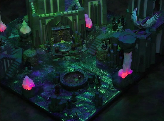 A battlemap of a walled courtyard under cover of night. Four towers stand in a row with a metal sacrificial platform bearing a pentagram sitting before a center tower. The white-winged and scorpion-tailed Tertia Tachonis is on top of a wall off the right-most tower. In front of the pentagram is a circular pool of dark red blood. Interspersed throughout the chamber are six red glowing crystal pillars. One ballista is on the lower platform of the left tower, and another is on top of the right tower. Pin, Vaelus, Julien, Occtis, Dr. Talter, and Thaisha stand at the edge of the battlemap, looking towards the pool of blood.