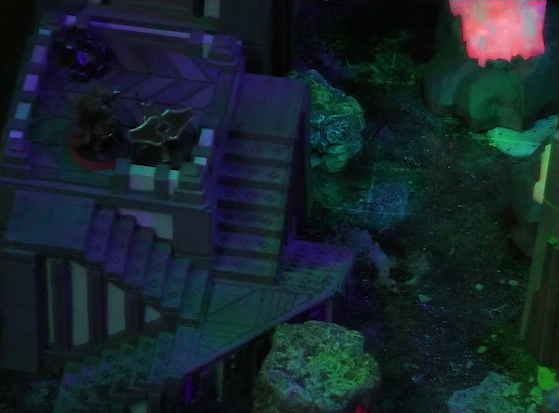 A night-time battlemap focused on the top of a square stone tower. Thaisha stands next to a ballista, pointing it off the corner of the tower’s roof. Two flights of stairs climb the tower. Off to the side is a red glowing crystal pillar.