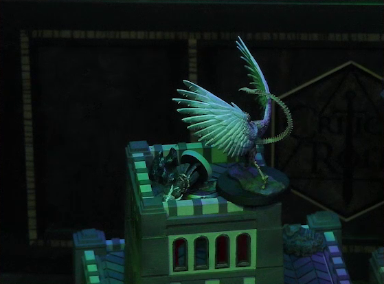 A night-time battlemap focused on the top of a square stone tower, where the white-winged and scorpion-tailed Tertia Tachonis stands over the body of Vaelus. Tertia leans forward with clawed hands, her wings outstretched and her skeletal tail arched above her head.