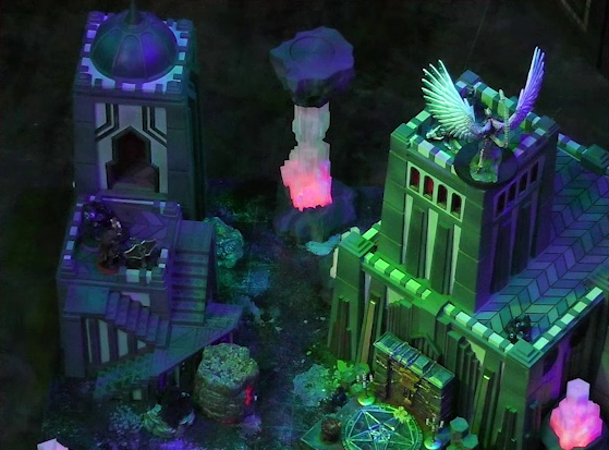 A battlemap of a walled courtyard under cover of night. The white-winged and scorpion-tailed Tertia Tachonis stands at the top of a tower over the body of Vaelus. Thaisha stands on a lower platform of a tower next to it, pointing a ballista at Tertia. Dr. Talter is by her side. At the base of Tertia’s tower is a metal sacrificial platform bearing a pentagram. Two red glowing crystal pillars stand to either side of the tower.