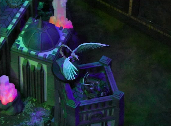 A battlemap of a walled courtyard under cover of night focused on the top of a tower. The white-winged and scorpion-tailed Tertia Tachonis stands at the edge of the tower over Julien. Beside Julien is a ballista. Next to the tower is another tower with a dome roof with a spike emerging from its center. Two red glowing crystal pillars rise from the ground.