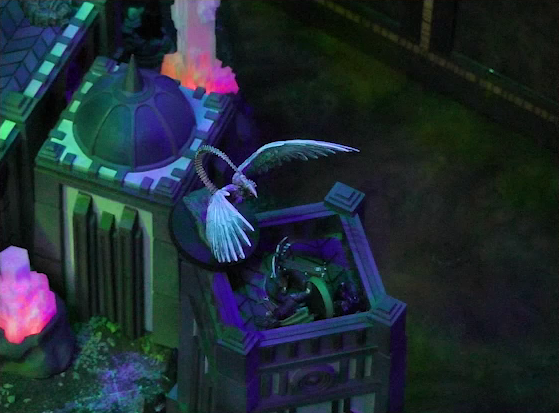 A battlemap of a walled courtyard under cover of night focused on the top of a tower. The white-winged and scorpion-tailed Tertia Tachonis stands at the edge of the tower over the body of Julien. Beside Julien is a ballista. Next to the tower is another tower with a dome roof with a spike emerging from its center. Two red glowing crystal pillars rise from the ground.