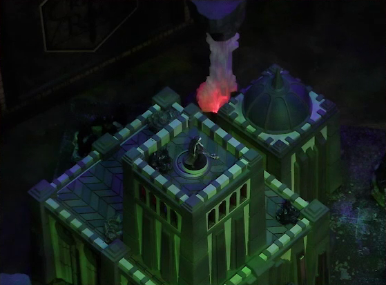 A battlemap of a walled courtyard under cover of night focused on Vaelus standing at the top of a tall square stone tower. Next to the tower is another tower with a dome roof with a spike emerging from its center. A red glowing crystal pillar rises in the background.