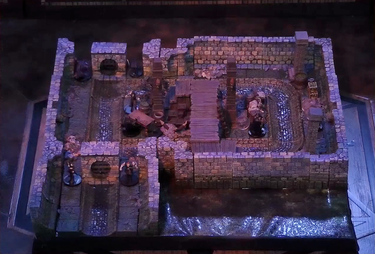 A battlemap of a stone underground room in the sewer. Five Crow Keepers stand within the room around a fallen Demodus Blix, while two guard an entrance on one side and two guard an entrance on the opposite side. The entrances have sewer grates set into them. A sewer trench runs in a circle in the room and exits through both entrances. A raised wooden boardwalk goes from the wall to the center of the room among wooden boxes and three stone pillars holding up the ceiling.