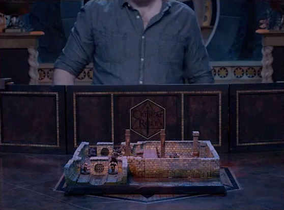 A battlemap of a stone underground room in the sewer, placed on the Critical Role table. Four Crow Keepers are visible guarding entrances on either side of the room, and three stone pillars rise up from the battlemap, intended to hold up the ceiling. The entrances have sewer grates set into them. Brennan is in the background behind a wooden DM screen, wearing a denim button-up with the top two buttons undone.