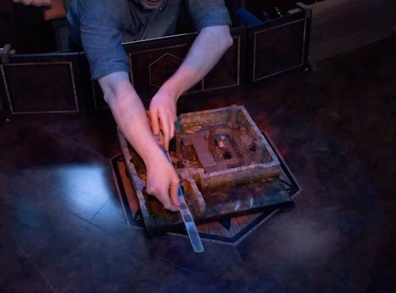 A battlemap of a stone underground room in the sewer, placed on the Critical Role table. Brennan holds a long transparent measuring tool over the battlemap, stretching out past the entrance closest to the camera. A few Crow Keepers are visible in the room not blocked by Brennan’s arms. A raised wooden boardwalk goes from the wall to the center of the room among wooden boxes and three stone pillars holding up the ceiling.