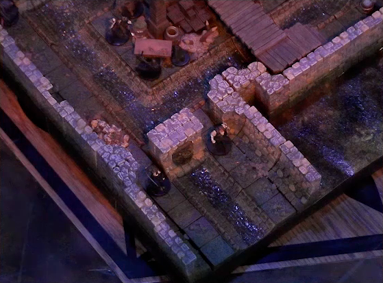 A battlemap of a stone underground room in the sewer, focused on an entrance guarded by two Crow Keepers. The entrance has a sewer grate set into it, and a sewer trench runs out the entrance from a circular channel within the room. In the room itself, three Crow Keepers stand by a number of wooden boxes, while another is visible guarding an entrance on the opposite side. A raised wooden boardwalk goes from the wall to the center of the room