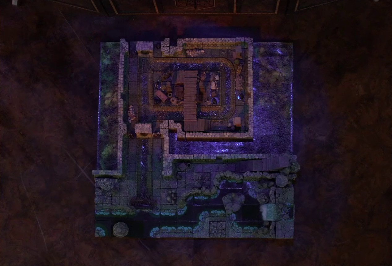 A top-down view of a battlemap of a stone underground room in the sewer. Five Crow Keepers and Laumy stand within the room around a fallen Demodus Blix, while two guard an entrance on the north side and two guard an entrance on the south side. The entrances have sewer grates set into them. A sewer trench runs in a circle in the room and exits through both entrances. A raised wooden boardwalk goes from the wall to the center of the room among wooden boxes and three stone pillars holding up the ceiling. An underground river runs past the room on the south end, and stairs lead down to a hallway between the river and the room.