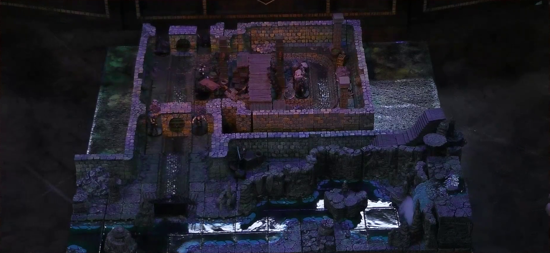 A battlemap of a stone underground room in the sewer. Bolaire hides around a corner from the room, down a hallway from a flight of stairs. Five Crow Keepers and Laumy stand within the room around a fallen Demodus Blix, while two guard an entrance on the north side and two guard an entrance on the south side. The entrances have sewer grates set into them. A sewer trench runs in a circle in the room and exits through both entrances. A raised wooden boardwalk goes from the wall to the center of the room among wooden boxes and three stone pillars holding up the ceiling. An underground river runs past the room on the south end.
