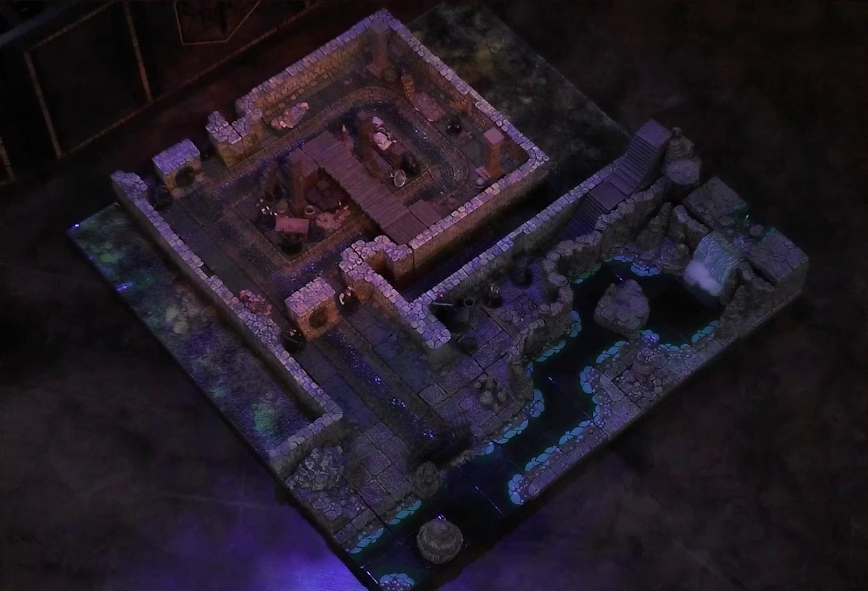 A battlemap of a stone underground room in the sewer. Bolaire hides around a corner from the room, down a hallway from a flight of stairs. Azune and Halandil are in the hallway with him. Five Crow Keepers and Laumy stand within the room around a fallen Demodus Blix, while two guard an entrance on the north side and two guard an entrance on the south side. The entrances have sewer grates set into them. A sewer trench runs in a circle in the room and exits through both entrances. A raised wooden boardwalk goes from the wall to the center of the room among wooden boxes and three stone pillars holding up the ceiling. An underground river runs past the room on the south end.