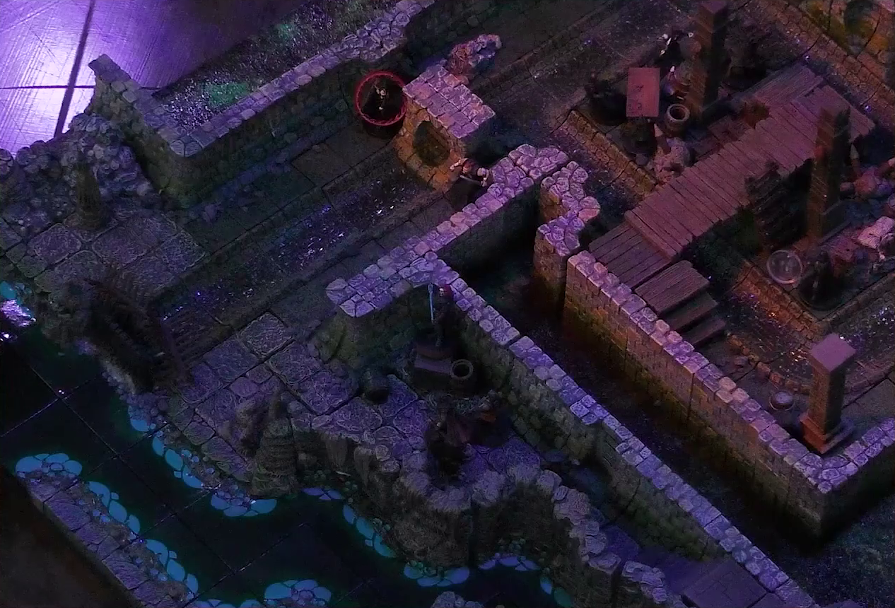 A battlemap of a stone underground room in the sewer. Bolaire hides around a corner from the room, down a hallway from a flight of stairs. Azune and Halandil are in the hallway with him. Five Crow Keepers and Laumy stand within the room around a fallen Demodus Blix, while two guard an entrance on the south side. One of them has a red status ring around his mini. The entrance has a sewer grate set into it. A sewer trench runs in a circle in the room and exits through the entrance. A raised wooden boardwalk goes from the wall to the center of the room among wooden boxes and three stone pillars holding up the ceiling. An underground river runs past the room on the south end.