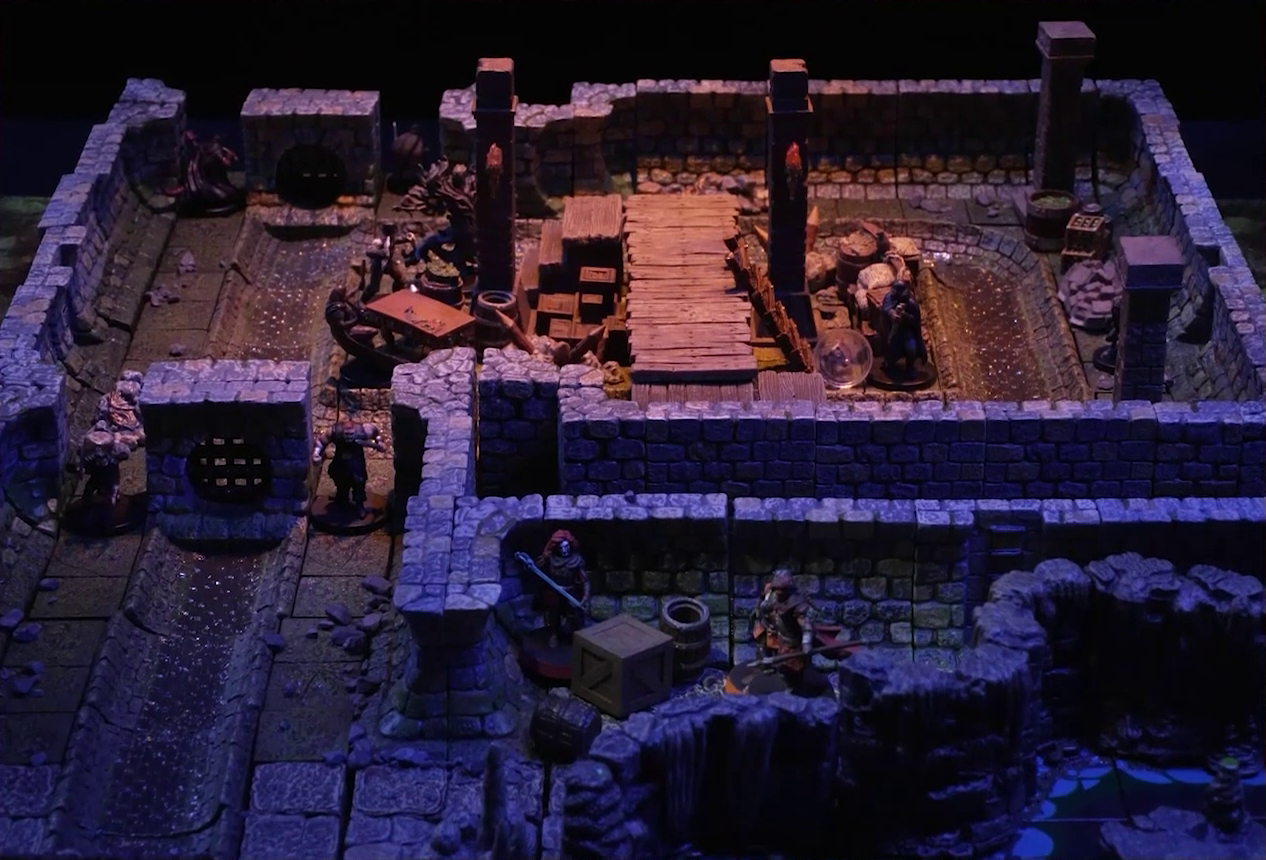 A battlemap of a stone underground room in the sewer. Bolaire hides around a corner from the room, down a hallway from a flight of stairs. Azune and Halandil are in the hallway with him. Five Crow Keepers and Laumy stand within the room around a fallen Demodus Blix, while two guard an entrance on the north side and two guard an entrance on the south side. The entrances have sewer grates set into them. A sewer trench runs in a circle in the room and exits through both entrances. A raised wooden boardwalk goes from the wall to the center of the room among wooden boxes and three stone pillars holding up the ceiling. An underground river runs past the room on the south end.