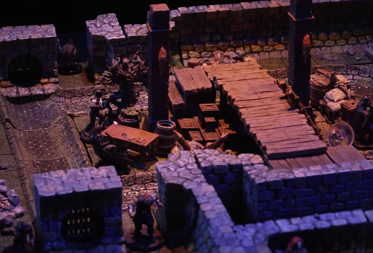 A battlemap of a stone underground room in the sewer. Laumy stands beside two Crow Keepers, while another stands over a fallen Demodus Blix a short distance away. Two other Crow Keepers guard an entrance on the north side and two guard an entrance on the south side. The entrances have sewer grates set into them. A sewer trench runs in a circle in the room and exits through both entrances. A raised wooden boardwalk goes from the wall to the center of the room among wooden boxes and three stone pillars holding up the ceiling.