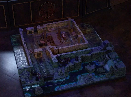A battlemap of a stone underground room in the sewer. Bolaire, Azune, Halandil, and Murray fight Crow Keepers in a hallway around the corner from the room. Crow Keepers and Laumy stand within the room around a fallen Demodus Blix, while one guards an entrance on the north side and none guard an entrance on the south side. The entrances have sewer grates set into them. A sewer trench runs in a circle in the room and exits through both entrances. A raised wooden boardwalk goes from the wall to the center of the room among wooden boxes and three stone pillars holding up the ceiling. An underground river runs past the room on the south end.
