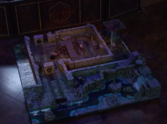 A battlemap of a stone underground room in the sewer. Bolaire, Azune, and Murray stand on a raised platform above the room indicating an upper floor. Halandil stands in a hallway below, outside the room. Crow Keepers and Laumy stand within the room around a fallen Demodus Blix, while one guards an entrance on the north side and none guard an entrance on the south side. The entrances have sewer grates set into them. A sewer trench runs in a circle in the room and exits through both entrances. A raised wooden boardwalk goes from the wall to the center of the room among wooden boxes and three stone pillars holding up the ceiling. An underground river runs past the room on the south end.