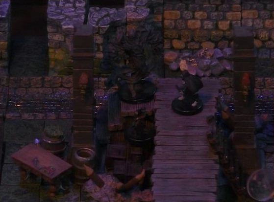 A battlemap of a stone underground room in the sewer, focused on Laumy and two Crow Keepers on a raised wooden boardwalk. Demodus Blix lies on the ground at the base of a ladder just at the edge of the frame. Two pillars rise on either side of the boardwalk with torches attached to them. A pile of boxes are on the ground next to the boardwalk.