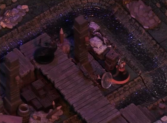 A battlemap of a stone underground room in the sewer, focused on Laumy on a raised wooden boardwalk, and Halandil standing over a fallen Demodus Blix on the ground next to the boardwalk. Two pillars rise on either side of the boardwalk with torches attached to them. A pile of boxes are on the ground next to the boardwalk. A sewer trench runs in a circle in the room.